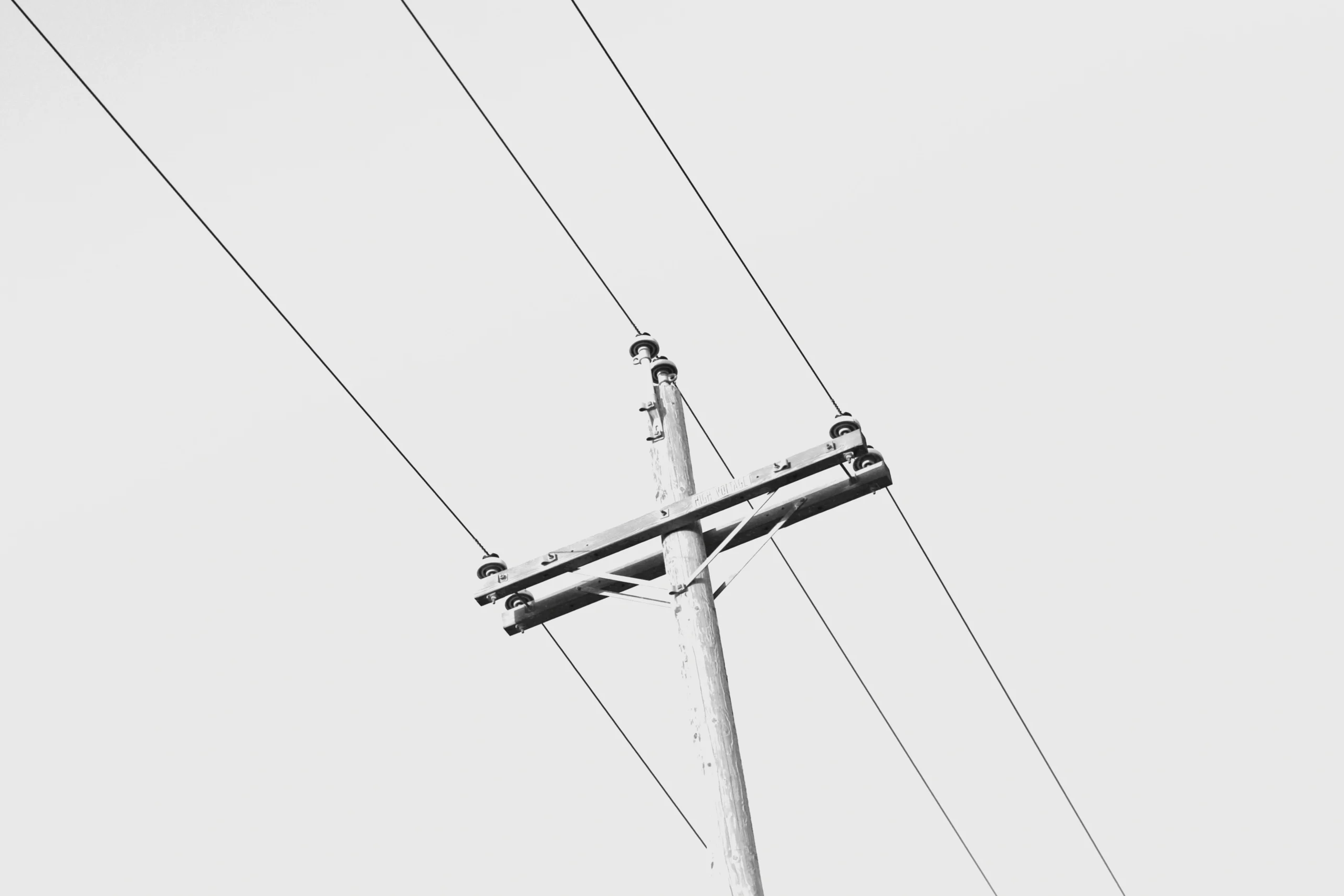Fotografia em preto e branco de um poste de madeira de rede elétrica, mostrando fios condutores de alta tensão sustentados por isoladores na cruzeta, contra um céu claro.