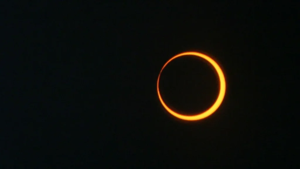 Fotografia em alto contraste de um eclipse solar anelar contra um fundo totalmente escuro. No centro, o disco negro da silhueta lunar oculta a maior parte do Sol, deixando visível apenas uma borda fina, perfeitamente circular e brilhante de luz alaranjada, formando o característico "anel de fogo".